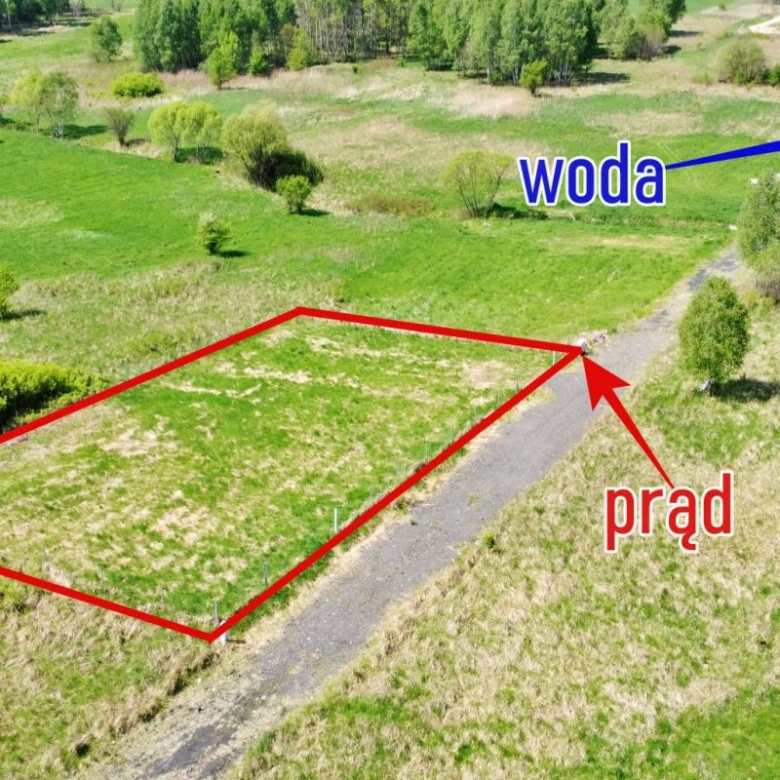 Aerial view of a rectangular plot outlined in red along a dirt road; the red arrow marks electricity access labeled 'prąd'; a blue arrow in the distance marks water labeled 'woda'.