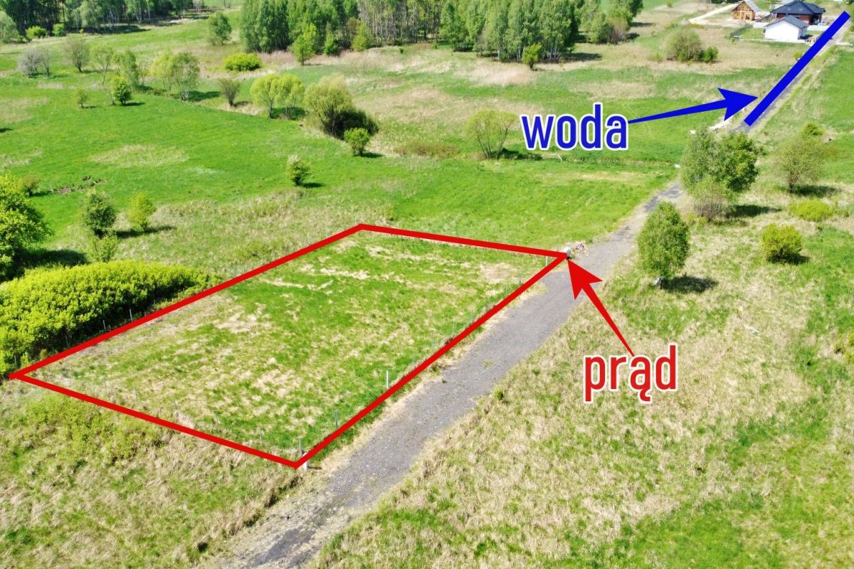 Aerial view of a rectangular plot outlined in red along a dirt road; the red arrow marks electricity access labeled 'prąd'; a blue arrow in the distance marks water labeled 'woda'.