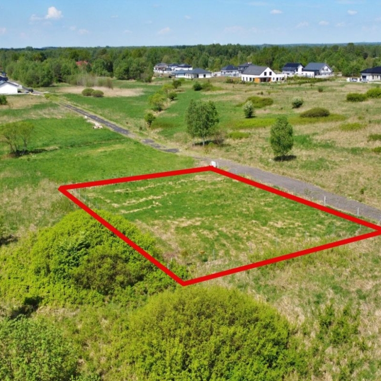 A vacant rectangular lot outlined in red within a grassy field, with a dirt road along one side and a few trees in the distance, in a developing residential area.