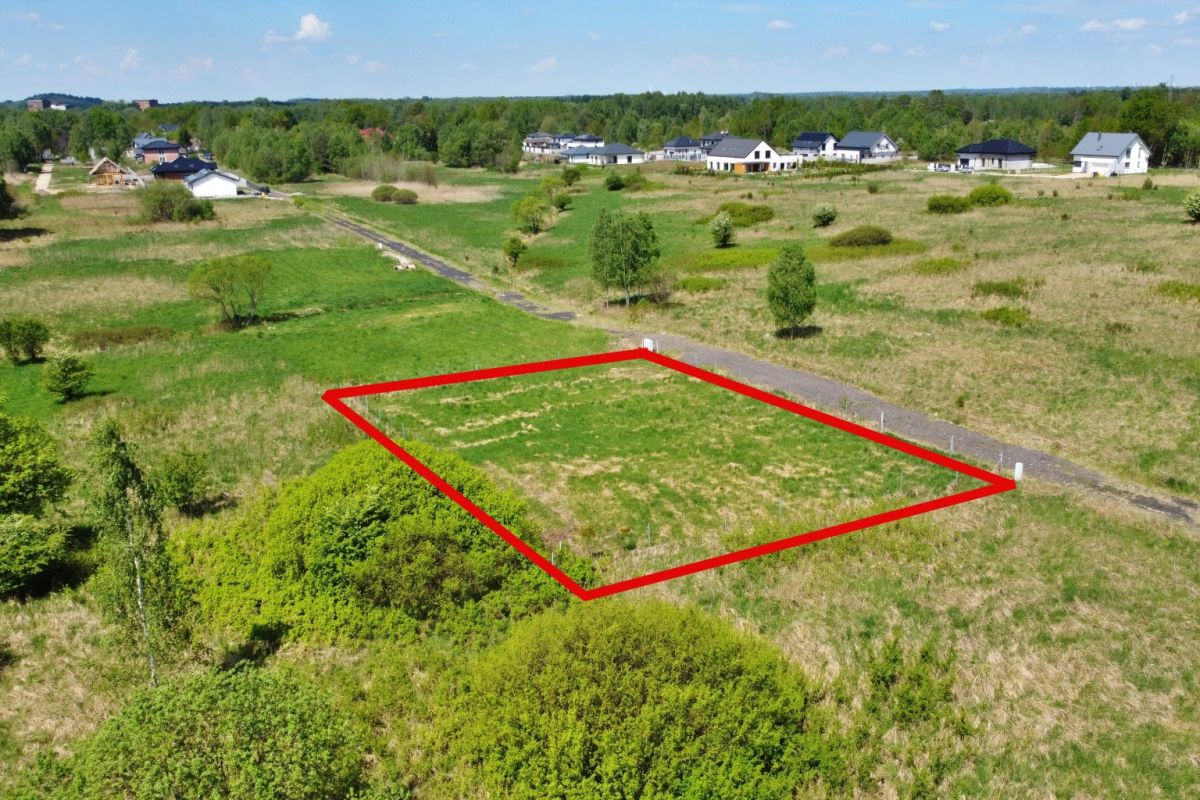 A vacant rectangular lot outlined in red within a grassy field, with a dirt road along one side and a few trees in the distance, in a developing residential area.