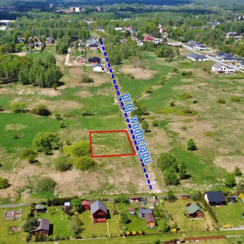 Aerial view of a vacant rectangular lot outlined in red in a grassy field, with a blue dashed line marking a nearby road and houses along the edge of the area.
