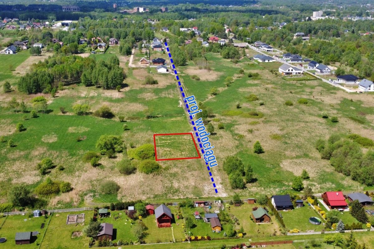 Aerial view of a vacant rectangular lot outlined in red in a grassy field, with a blue dashed line marking a nearby road and houses along the edge of the area.