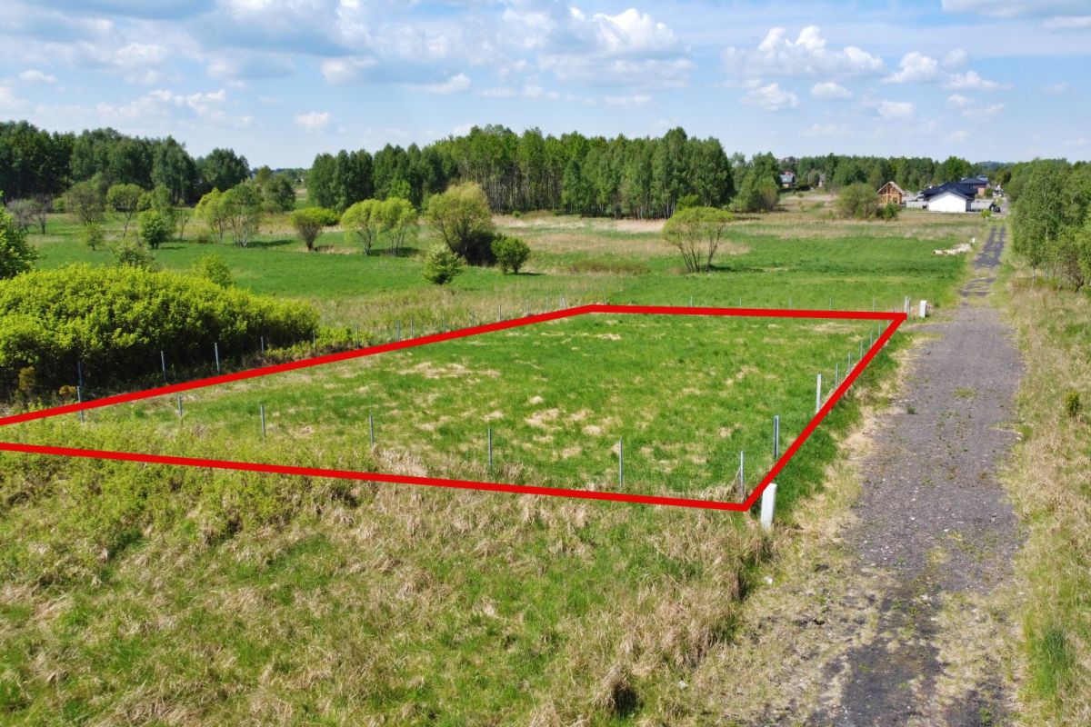 Rectangular grassy lot bordered by red boundary lines and fence posts, beside a gravel rural road.