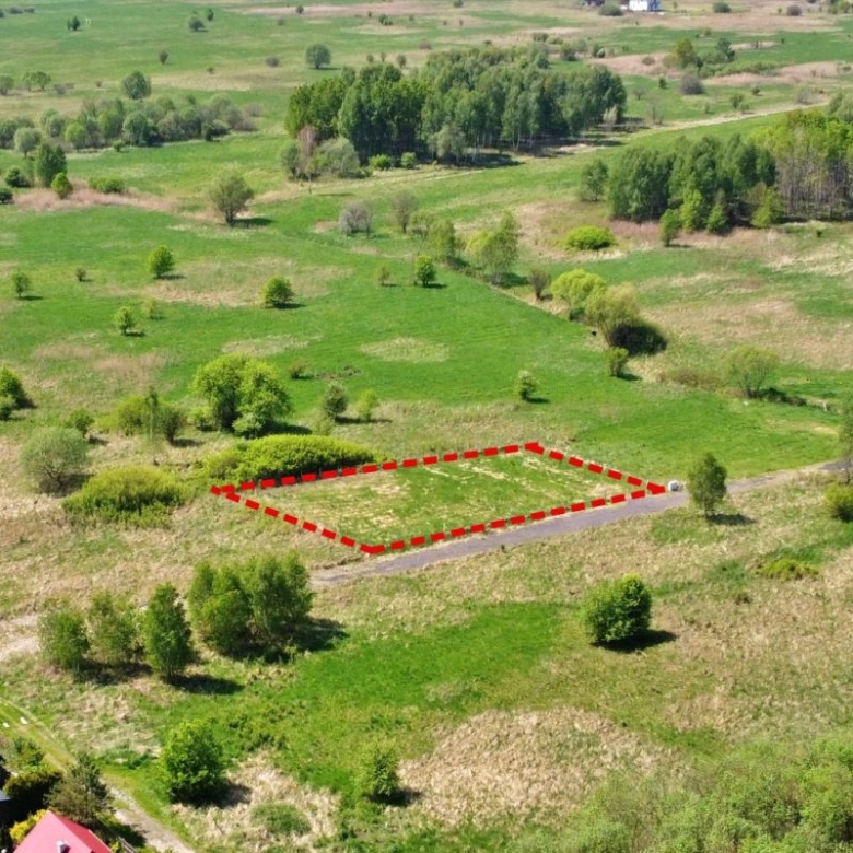 A vacant rectangular plot outlined in red dashed lines beside a country road, surrounded by green fields and trees.