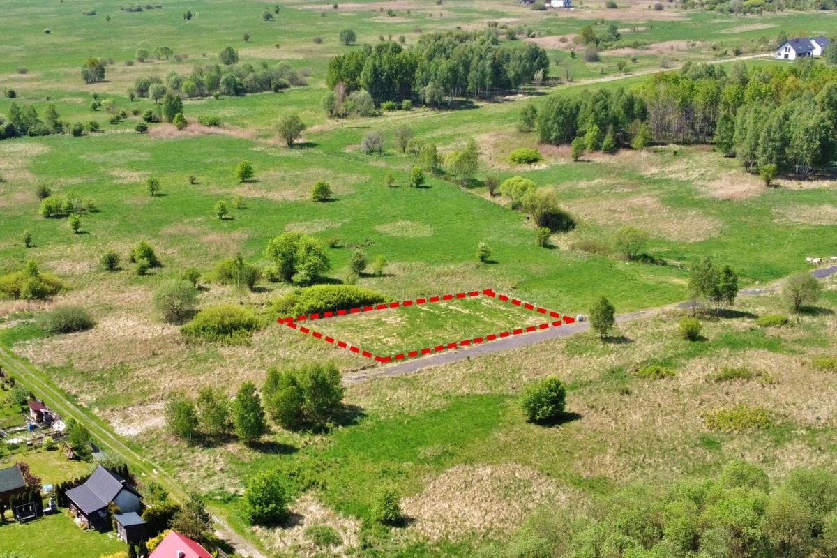 A vacant rectangular plot outlined in red dashed lines beside a country road, surrounded by green fields and trees.