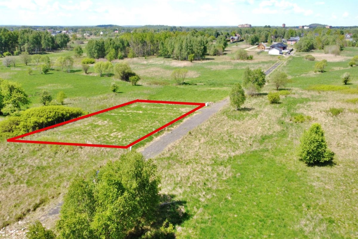 Outline of a rectangular plot of land marked in red along a rural road, surrounded by green fields and scattered trees for sale or development.