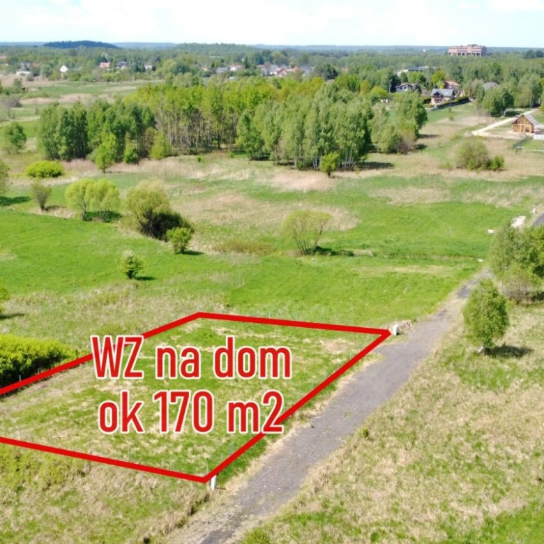 Aerial view of a vacant plot outlined in red for a house, labeled 'WZ na dom ok 170 m2' with a gravel road nearby and green landscape surrounding it.