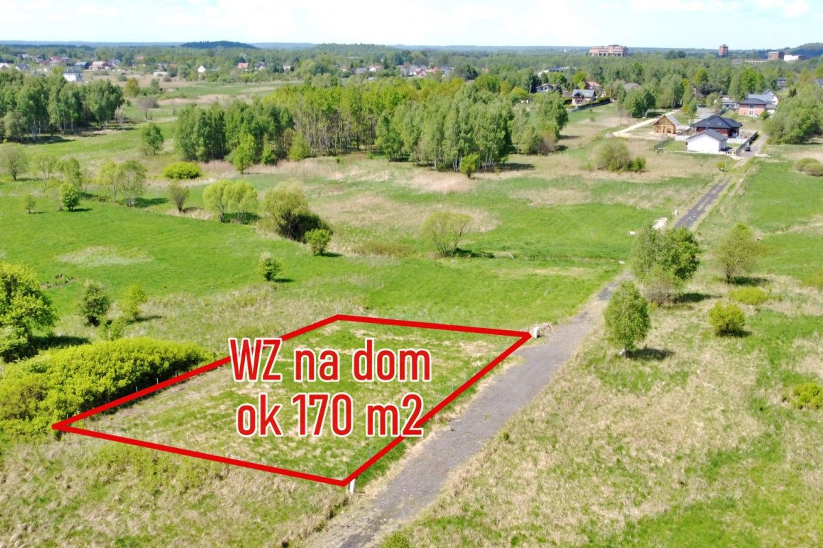Aerial view of a vacant plot outlined in red for a house, labeled 'WZ na dom ok 170 m2' with a gravel road nearby and green landscape surrounding it.
