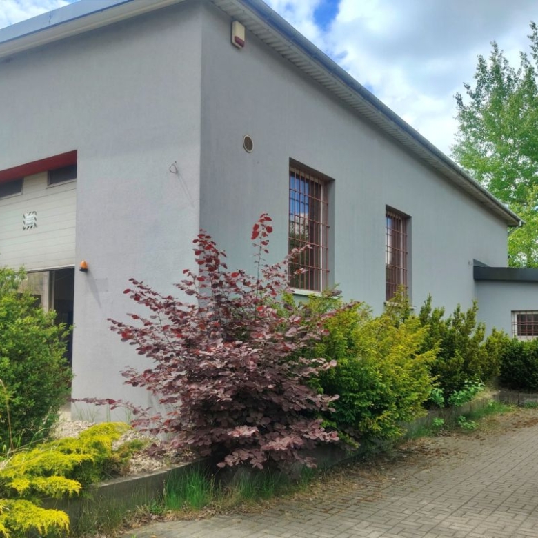 Exterior of a gray industrial building with red trim, two barred windows, and a paved walkway bordered by green shrubs and a burgundy plant bed.