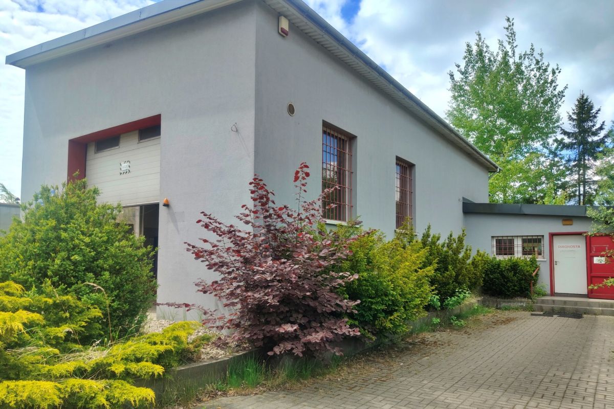 Exterior of a gray industrial building with red trim, two barred windows, and a paved walkway bordered by green shrubs and a burgundy plant bed.
