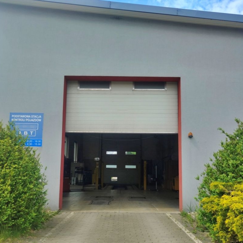 Open vehicle inspection bay inside a gray building framed by red border, with a blue sign on the left and green shrubs nearby.