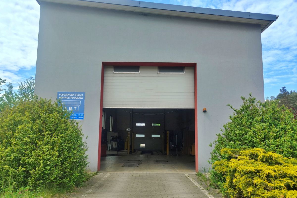Open vehicle inspection bay inside a gray building framed by red border, with a blue sign on the left and green shrubs nearby.