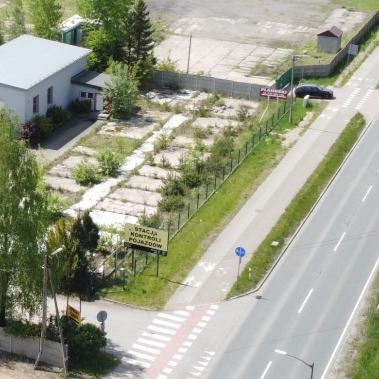 Aerial view of a small vehicle inspection station lot beside a main road, with a beige building, fenced lot, and a crosswalk nearby.