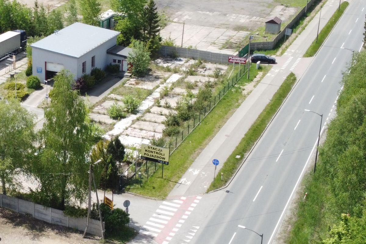 Aerial view of a small vehicle inspection station lot beside a main road, with a beige building, fenced lot, and a crosswalk nearby.