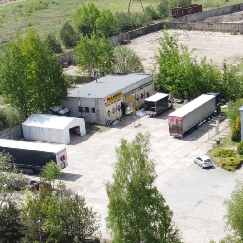 Aerial view of a truck loading yard with several semi-trailers and a gray warehouse, surrounded by trees and greenery; a white car sits near a blue-roofed building at right.