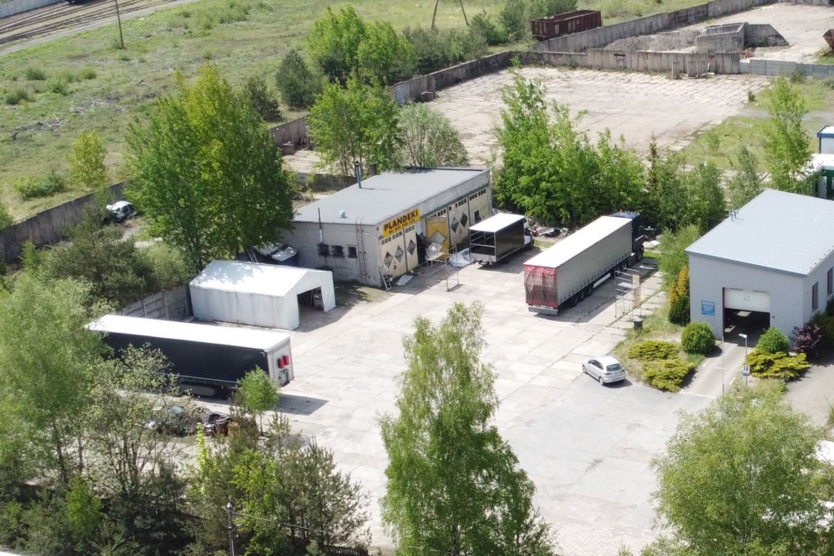 Aerial view of a truck loading yard with several semi-trailers and a gray warehouse, surrounded by trees and greenery; a white car sits near a blue-roofed building at right.