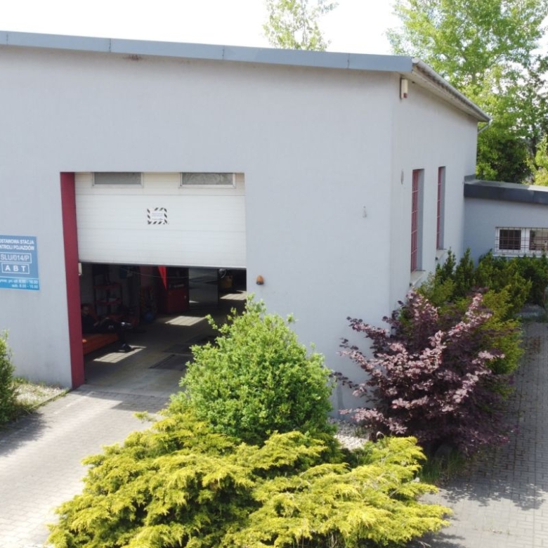 Industrial workshop with a large open garage bay; cars inside, light-gray building, surrounded by bushes and paved driveway