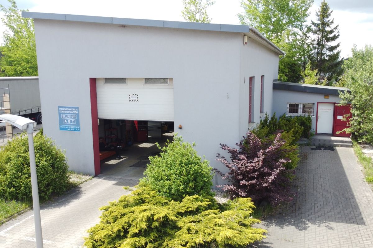 Industrial workshop with a large open garage bay; cars inside, light-gray building, surrounded by bushes and paved driveway