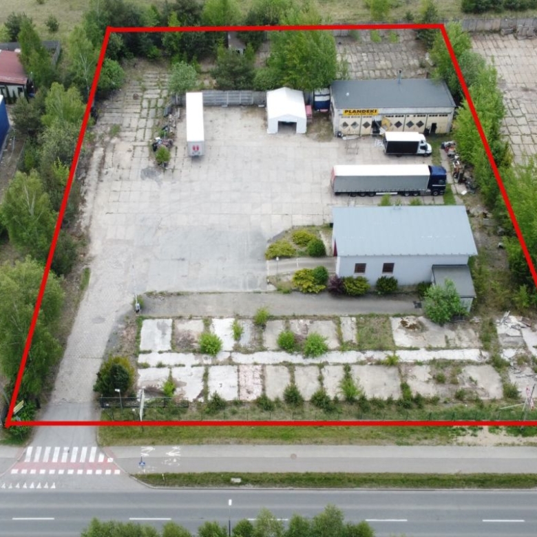 Aerial view of a fenced industrial yard outlined in red, with buildings, a truck trailer, and surrounding trees and roads.