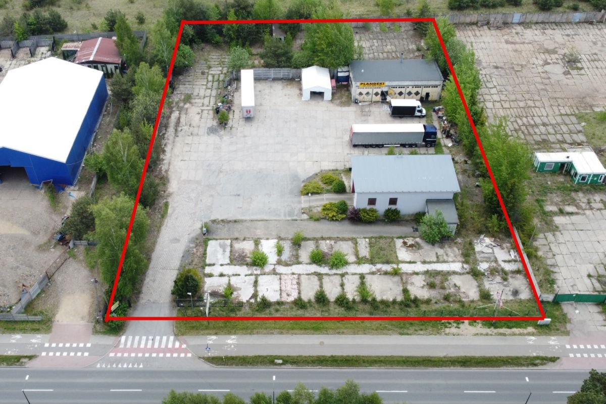 Aerial view of a fenced industrial yard outlined in red, with buildings, a truck trailer, and surrounding trees and roads.