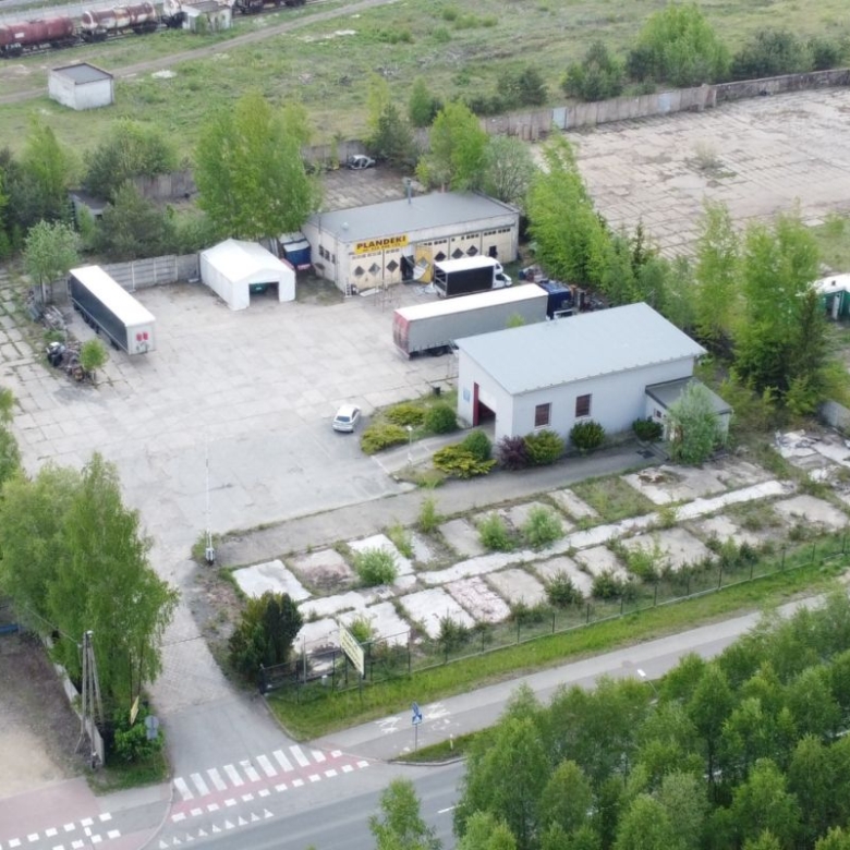 Aerial view of an industrial yard with small buildings, loading trailers, and fenced lots lined by trees near a road intersection.