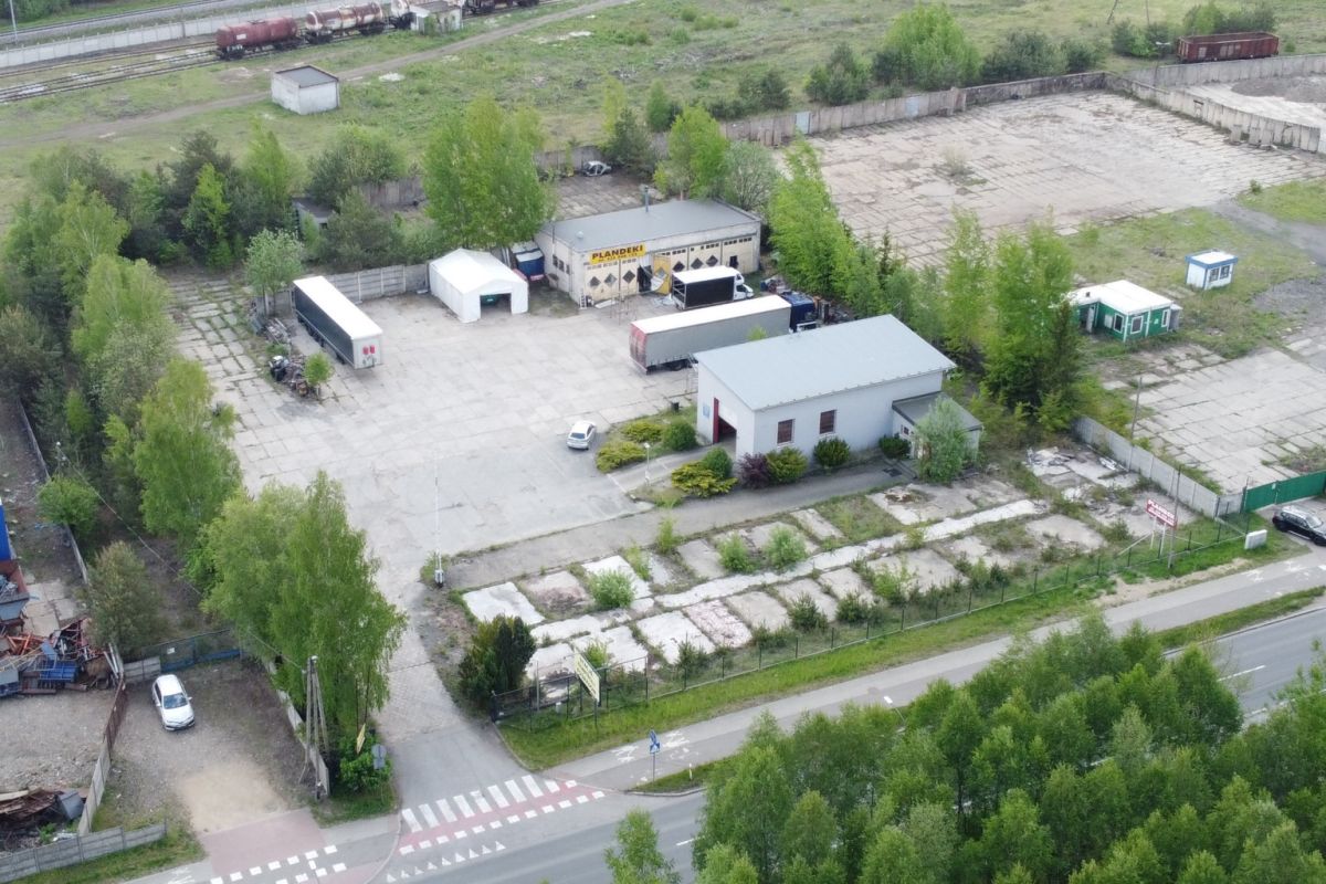 Aerial view of an industrial yard with small buildings, loading trailers, and fenced lots lined by trees near a road intersection.