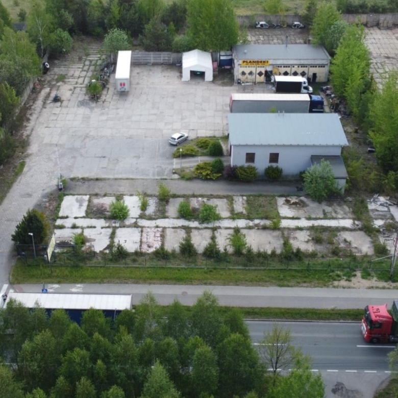 Aerial view of an industrial yard with small buildings, parked trucks, and a tree-lined perimeter along a main road.