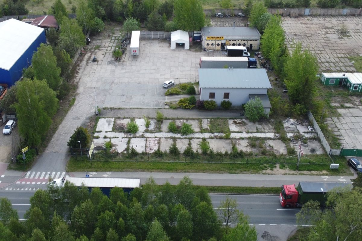 Aerial view of an industrial yard with small buildings, parked trucks, and a tree-lined perimeter along a main road.