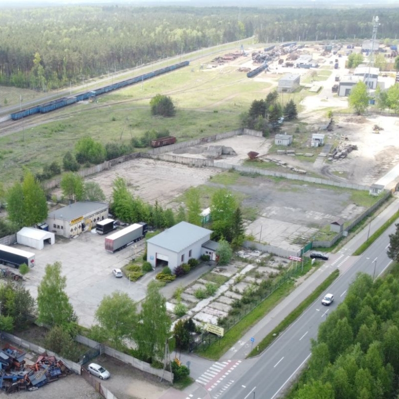 Aerial view of an industrial yard with warehouses, parked trucks, and a tree-lined road nearby in a green landscape.|Aerial view shows railway tracks with blue freight cars beside an industrial complex and open lots.|Large outdoor storage area with trucks, trailers, and scattered equipment near a road bordered by trees.|Industrial park from above: warehouses, loading bays, trucks, and a perimeter road with forest beyond.|Overhead shot of a logistics yard and rail line, with wooded areas on one side and distant buildings.