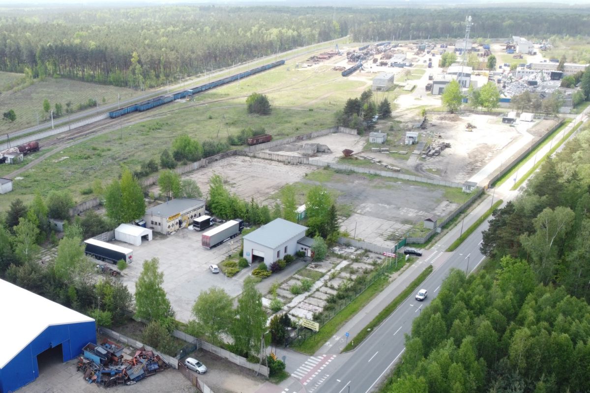 Aerial view of an industrial yard with warehouses, parked trucks, and a tree-lined road nearby in a green landscape.|Aerial view shows railway tracks with blue freight cars beside an industrial complex and open lots.|Large outdoor storage area with trucks, trailers, and scattered equipment near a road bordered by trees.|Industrial park from above: warehouses, loading bays, trucks, and a perimeter road with forest beyond.|Overhead shot of a logistics yard and rail line, with wooded areas on one side and distant buildings.