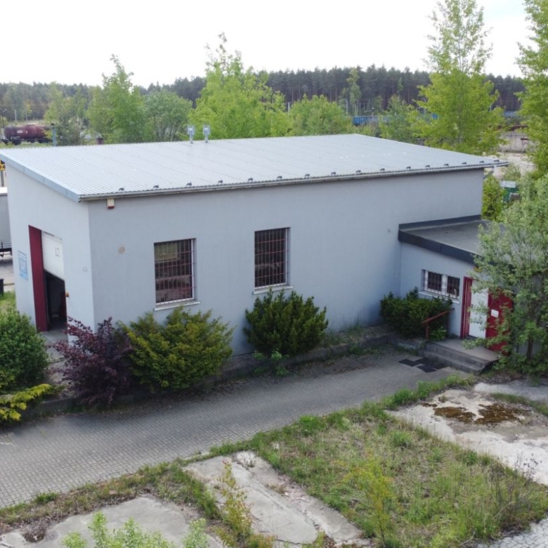 Light gray industrial warehouse with a metal roof, red door, and shrubs in a paved outdoor lot.