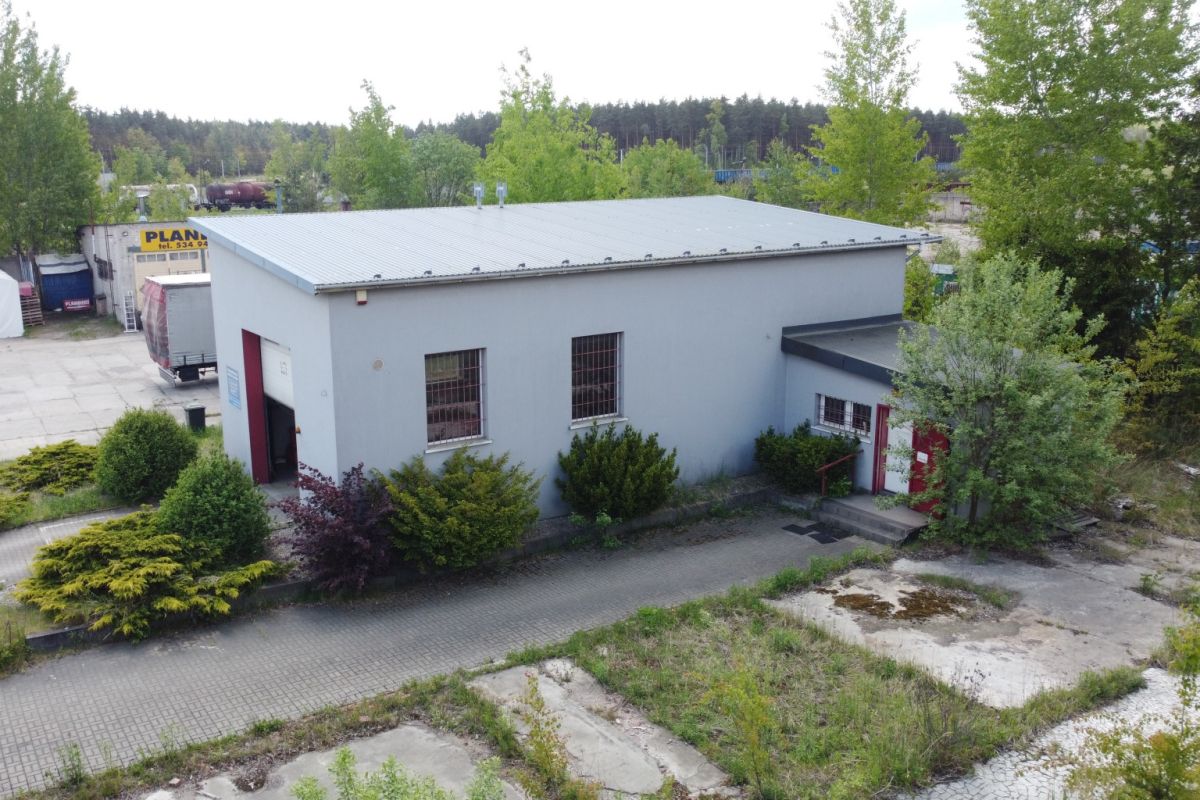 Light gray industrial warehouse with a metal roof, red door, and shrubs in a paved outdoor lot.