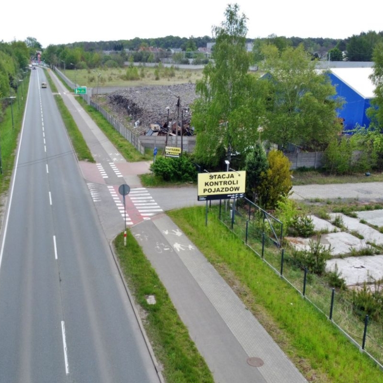 Aerial view of a two-lane road with a separate bike lane and pedestrian path, bordered by greenery and industrial buildings; a yellow sign reads "Stacja Kontroli Pojazdów" near a gated lot.