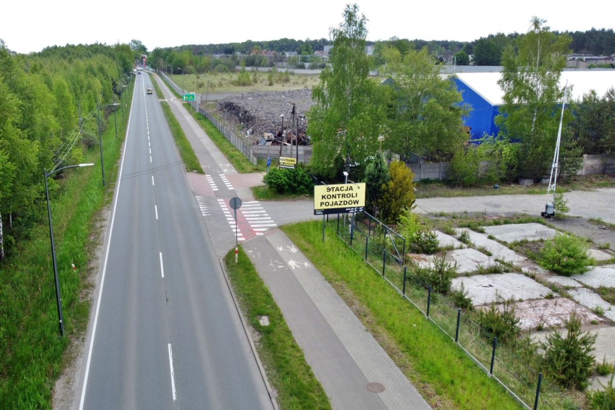 Aerial view of a two-lane road with a separate bike lane and pedestrian path, bordered by greenery and industrial buildings; a yellow sign reads "Stacja Kontroli Pojazdów" near a gated lot.