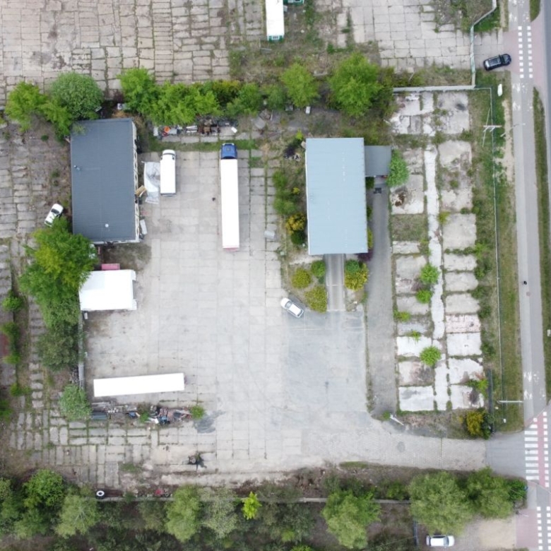 Aerial view of an industrial yard with two buildings, parked trucks, and trees along a road on the right side.
