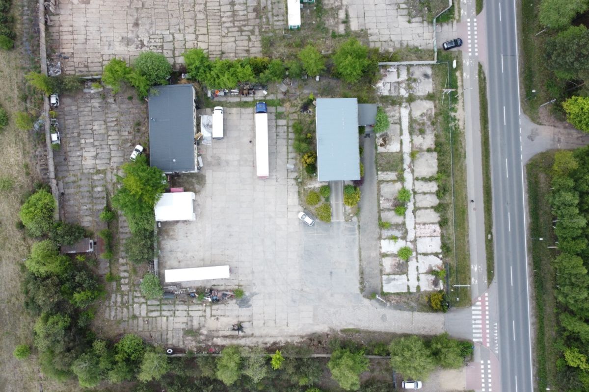 Aerial view of an industrial yard with two buildings, parked trucks, and trees along a road on the right side.
