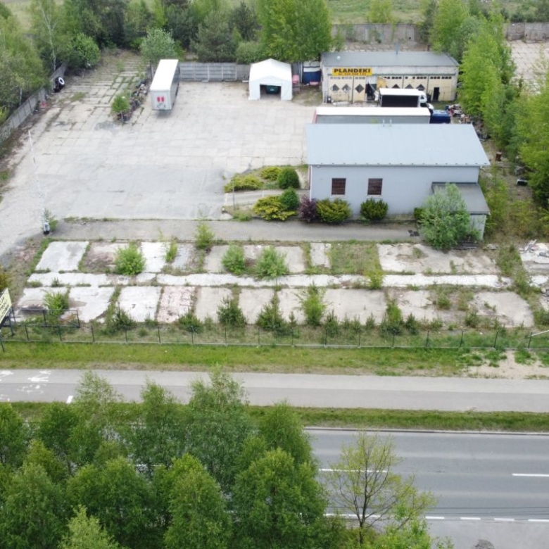 Aerial view of an industrial yard with a small grey building, vacant cracked concrete plots, and surrounding trees near a busy road at the bottom.