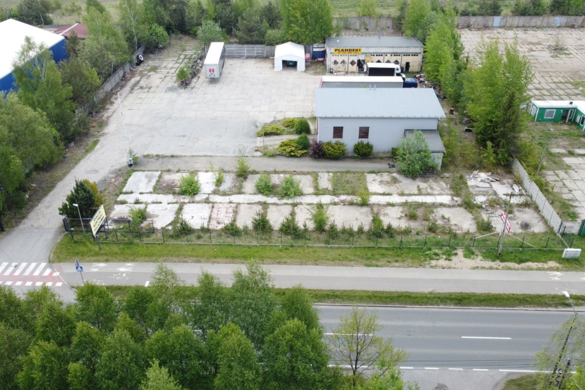 Aerial view of an industrial yard with a small grey building, vacant cracked concrete plots, and surrounding trees near a busy road at the bottom.
