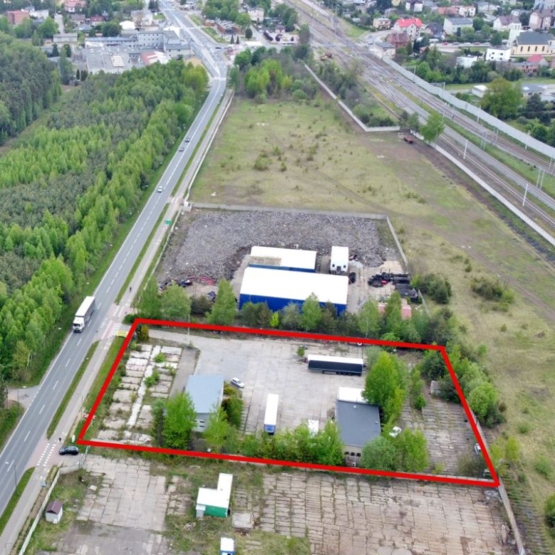 Aerial view of a fenced industrial yard outlined in red, containing small buildings, vehicles, and greenery beside a main road and railway tracks in the distance.