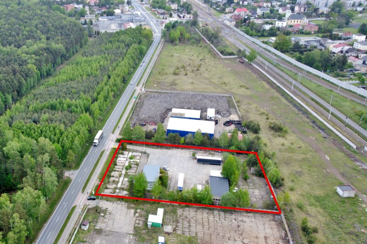 Aerial view of a fenced industrial yard outlined in red, containing small buildings, vehicles, and greenery beside a main road and railway tracks in the distance.