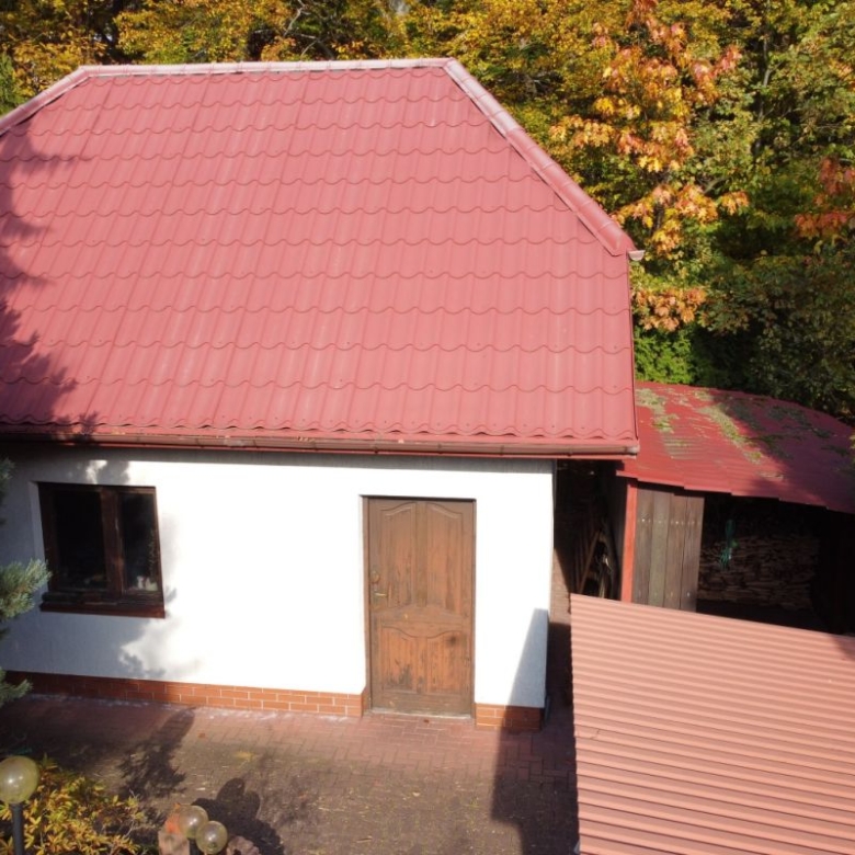 White single-story house with a large red metal roof, a wooden door and a small window, surrounded by autumn trees and greenery.