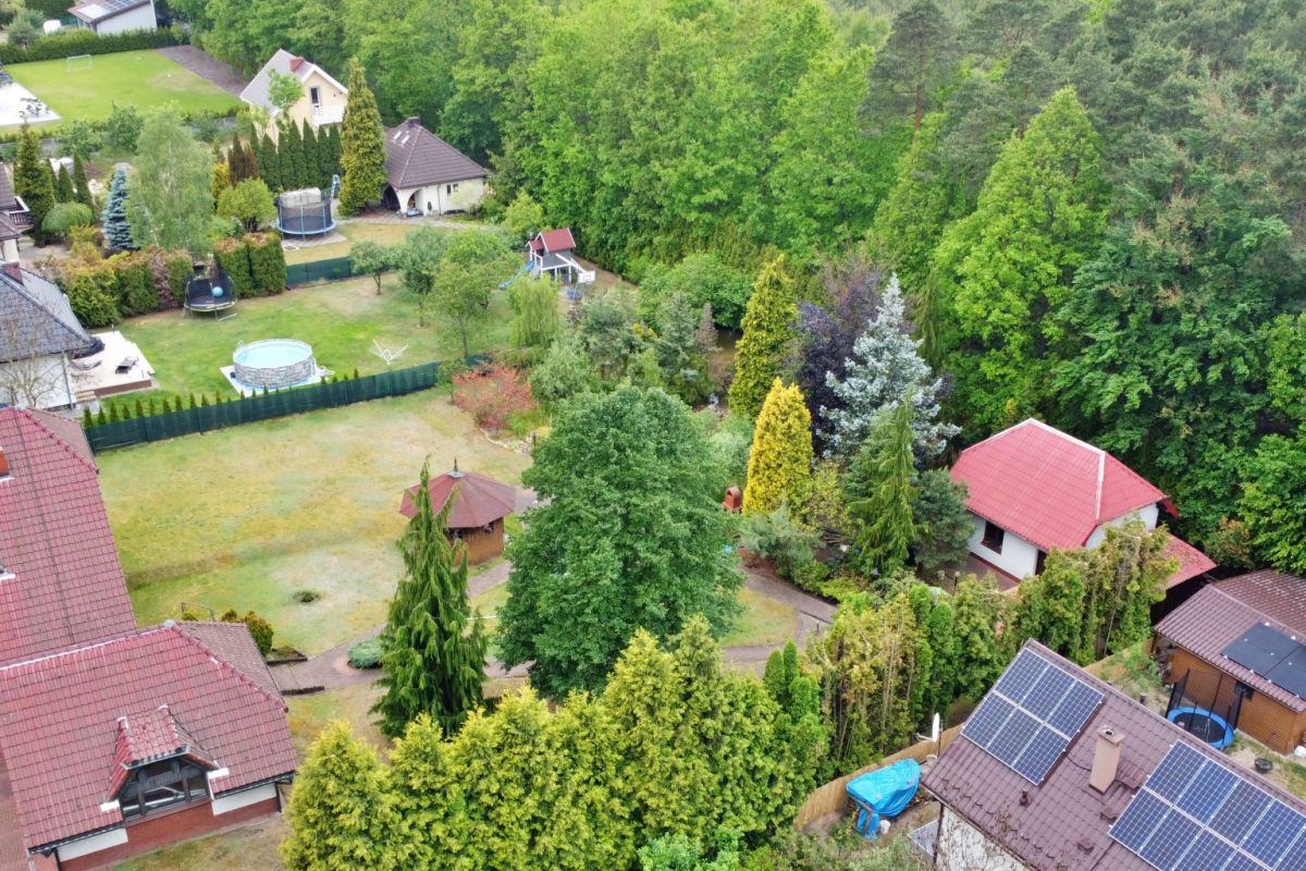 Aerial view of a suburban neighborhood with green lawns, trees, and several houses; solar panels on a roof in the foreground and a fenced garden area with a trampoline visible in the back yard.