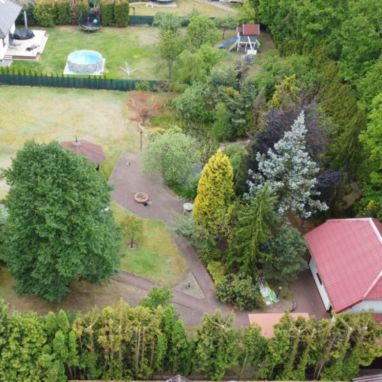 Aerial view of a backyard with a circular patio and fire pit surrounded by trees, a winding brick path, and a red-roofed outbuilding; a trampoline, pool, and play equipment visible in the distance.