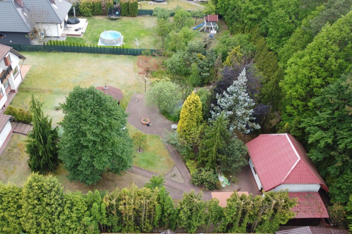 Aerial view of a backyard with a circular patio and fire pit surrounded by trees, a winding brick path, and a red-roofed outbuilding; a trampoline, pool, and play equipment visible in the distance.