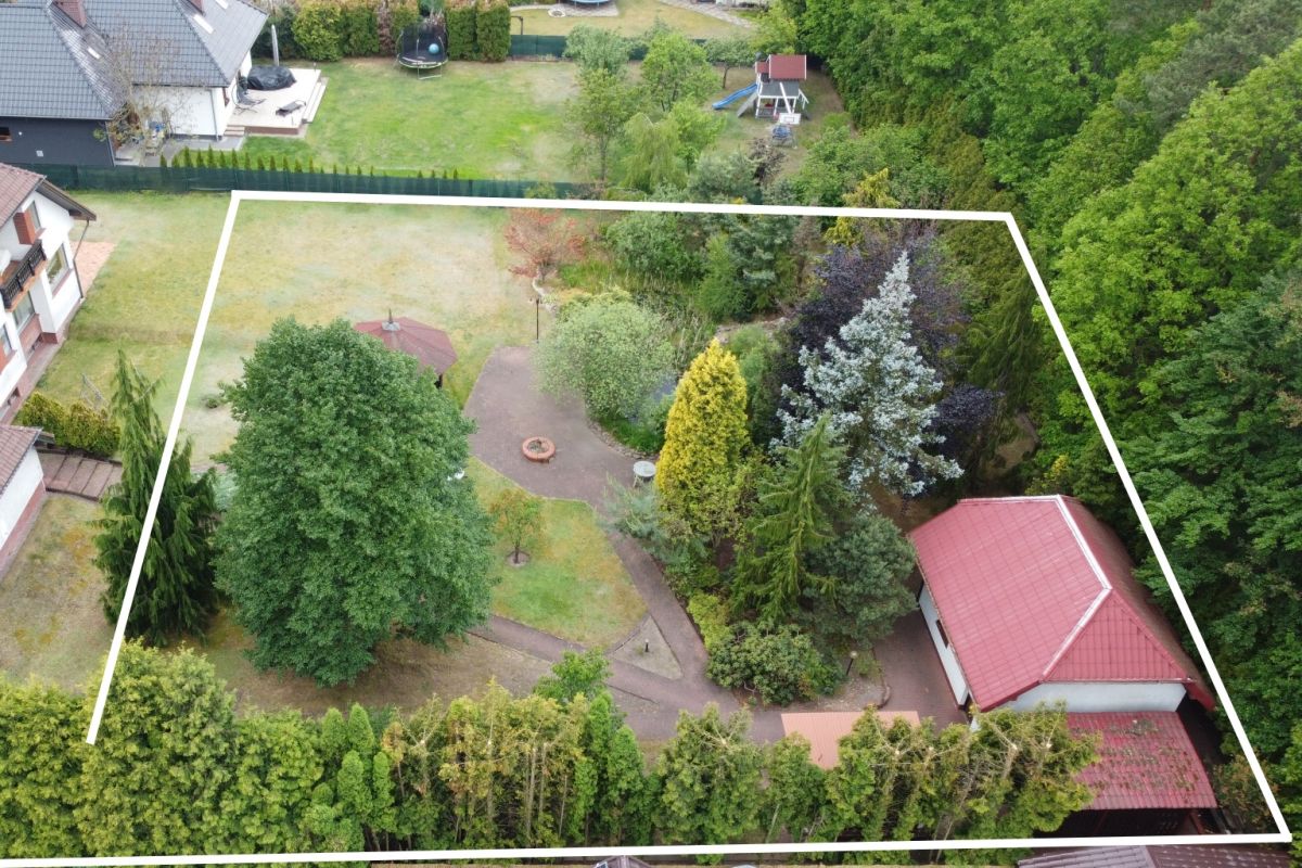Aerial view of a fenced backyard with trees, a circular patio, a fire pit, and a red-roofed shed surrounded by greenery.