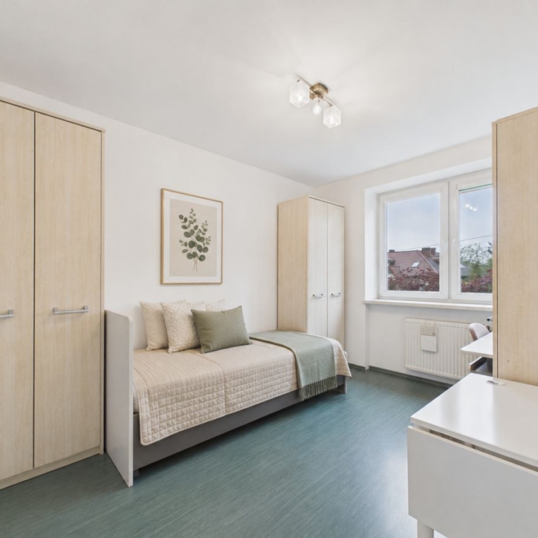 Minimal bedroom with light wood wardrobes, a single bed with beige quilt and green throw pillow, and a white desk near a window.