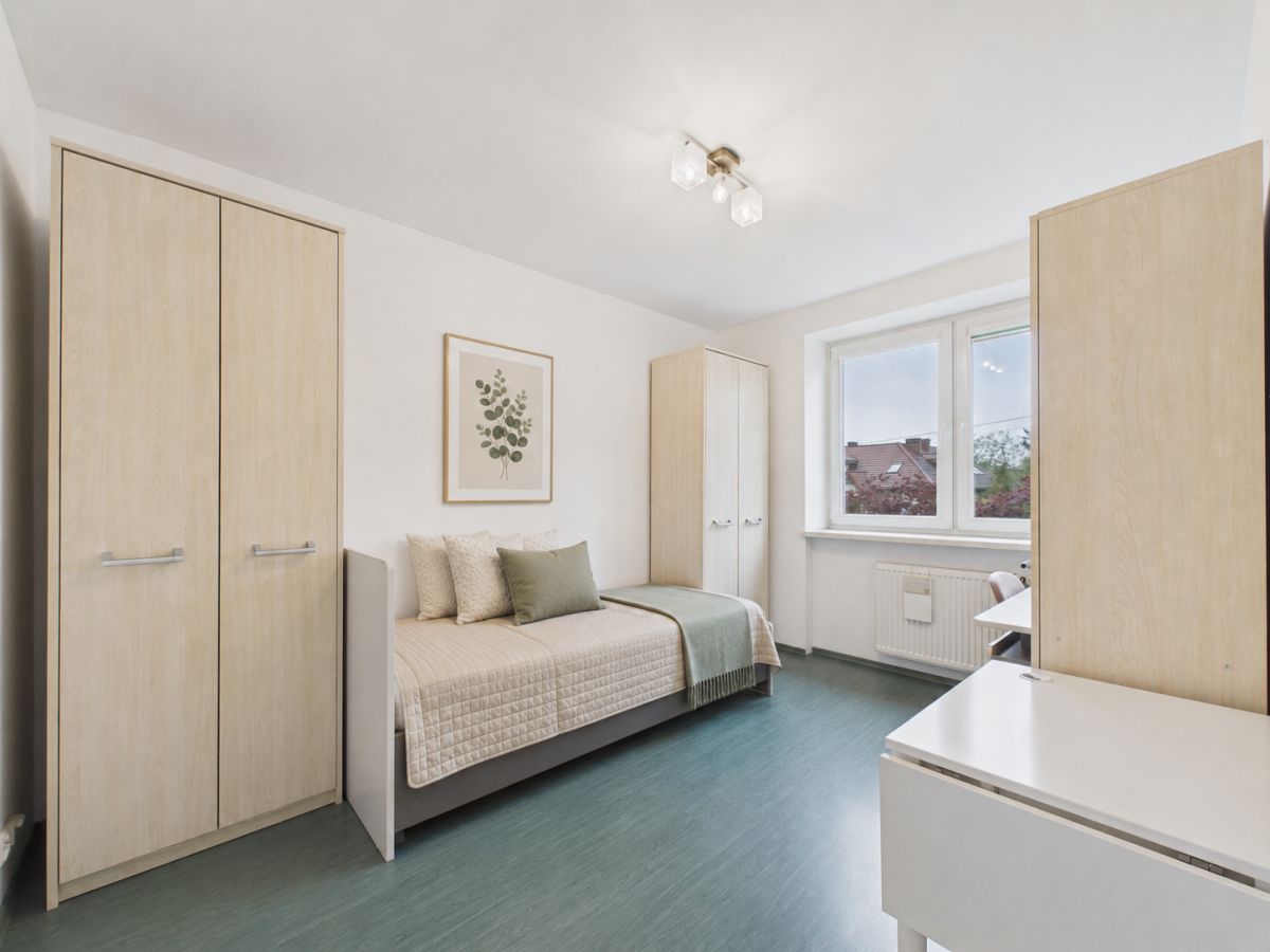 Minimal bedroom with light wood wardrobes, a single bed with beige quilt and green throw pillow, and a white desk near a window.
