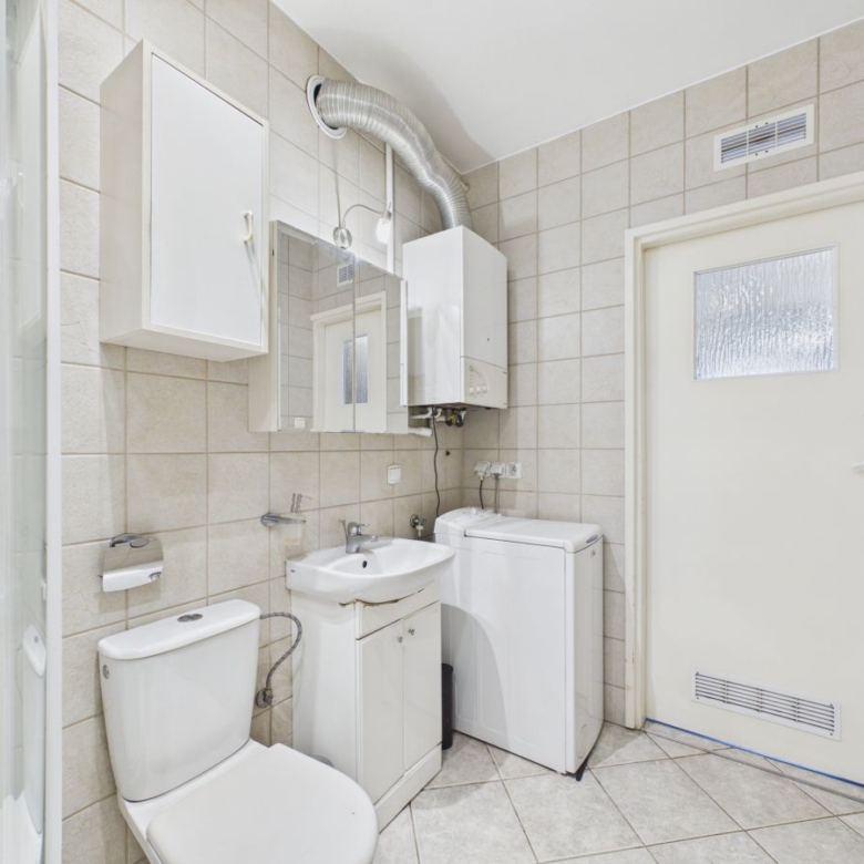 Small bathroom with toilet, sink and washing machine against beige tiled walls. A wall-mounted boiler and mirror cabinet are visible above the sink.