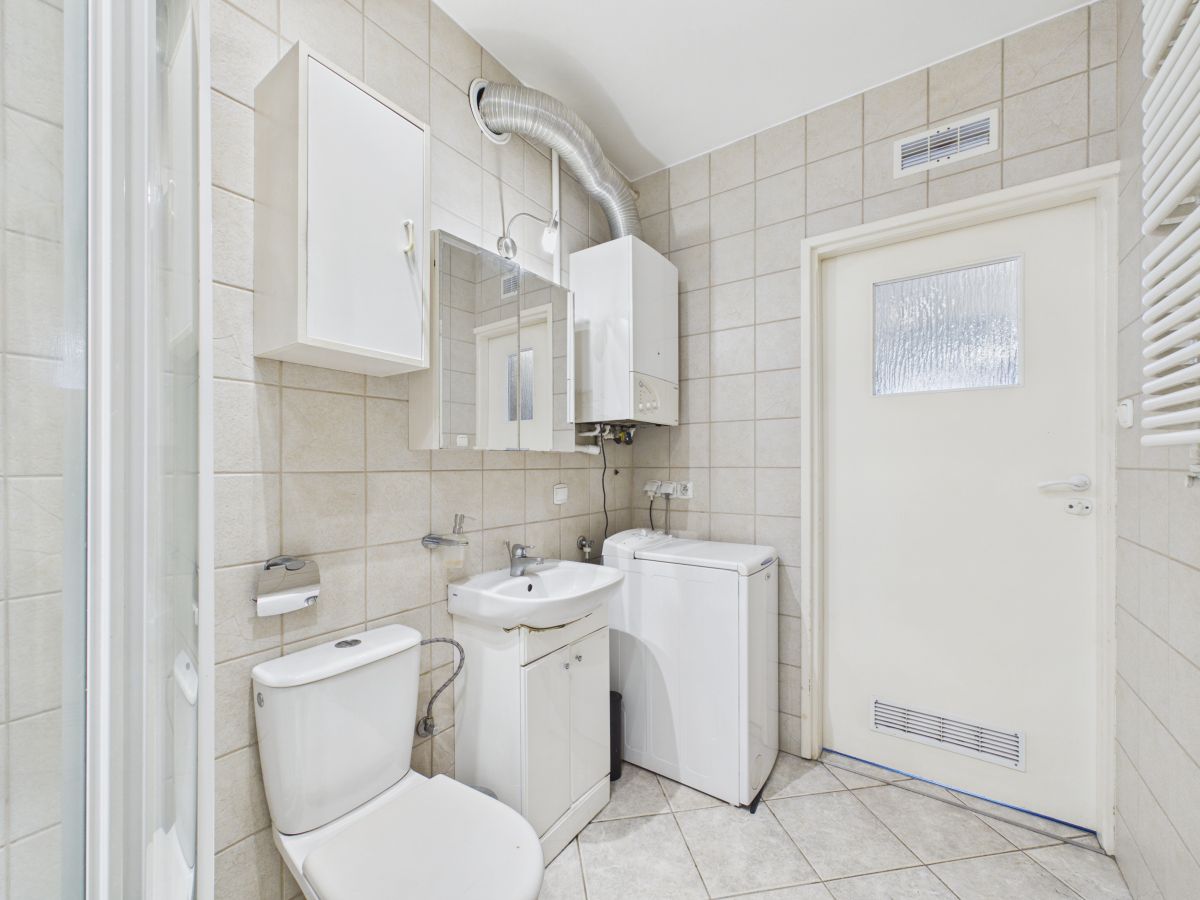 Small bathroom with toilet, sink and washing machine against beige tiled walls. A wall-mounted boiler and mirror cabinet are visible above the sink.