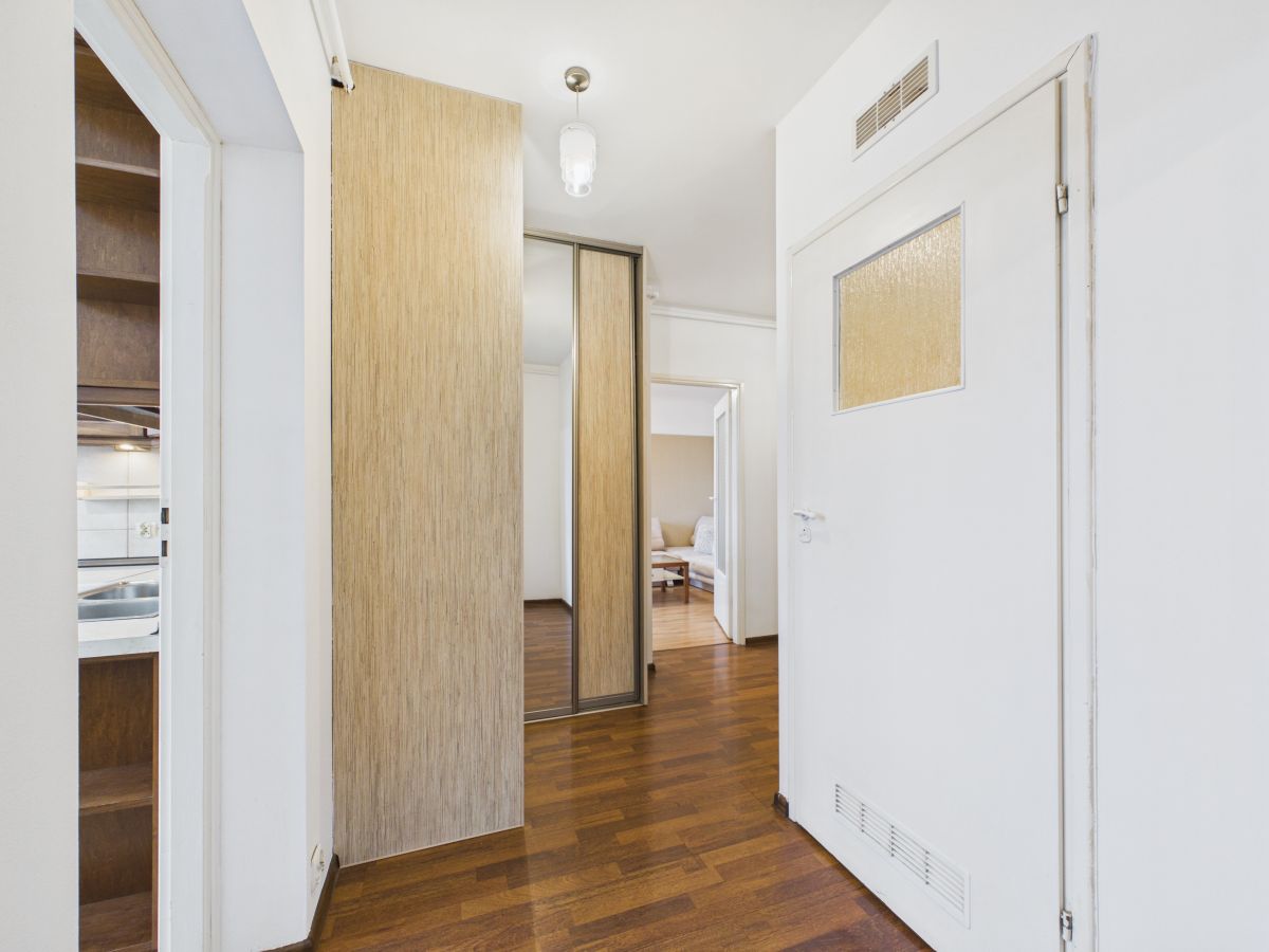 Bright hallway with wood flooring, a built-in wooden closet with mirrored sliding doors on the left, and a white door with a frosted window on the right; living room visible at the end.